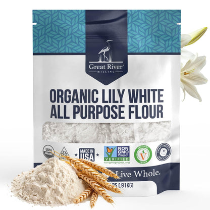 A 25 lb bag of Great River Milling Organic Lily White All-Purpose Flour features a blue patterned top and white base with clear labeling. Wheat stalks and a lily flower are pictured beside the bag