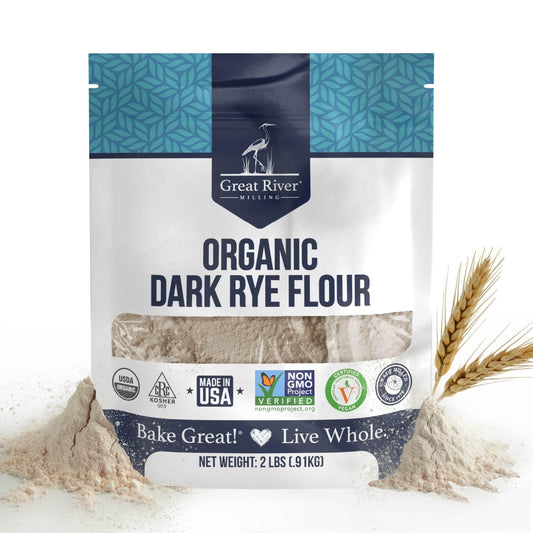 A 25 lb bag of Great River Milling Organic Dark Rye Flour with a blue geometric pattern on top, surrounded by scattered flour and wheat stalks. The package displays organic and non-GMO certifications