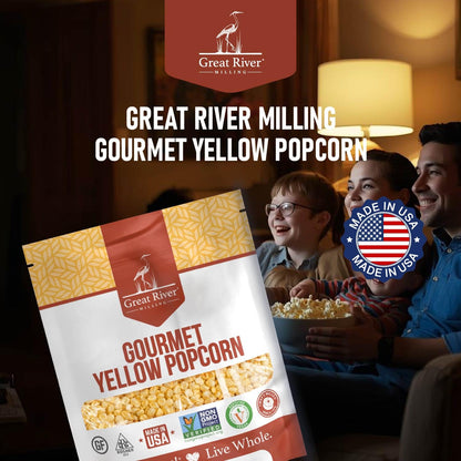 A bag of Great River Milling Gourmet Yellow Popcorn Kernels with a patterned yellow design and clear window, set against a cozy background with people enjoying popcorn