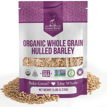 A 25 lb bag of Great River Milling Organic Hulled Barley with a purple and white design, featuring various certification logos. Loose barley grains surround the bag