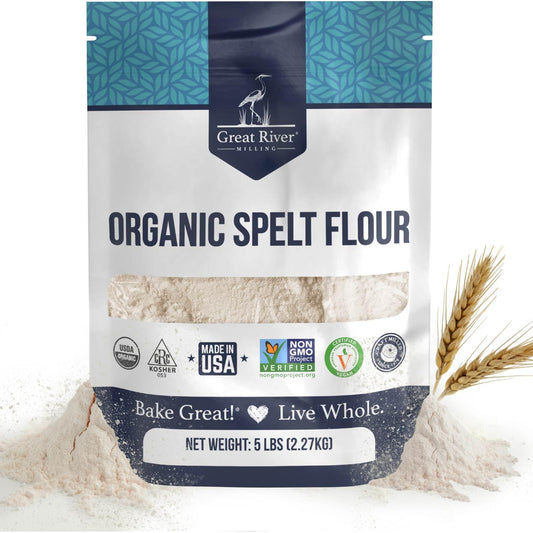 A 5 lb bag of Organic Spelt Flour, an ancient wheat flour from Great River Milling, sits beside wheat stalks, displaying various certification logos and a clear window that reveals the flour inside.