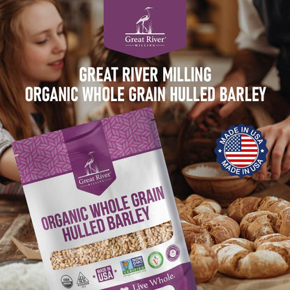 A 25 lb bag of Great River Milling Organic Hulled Barley is displayed with a purple and white design, surrounded by fresh bread and a "Made in USA" emblem