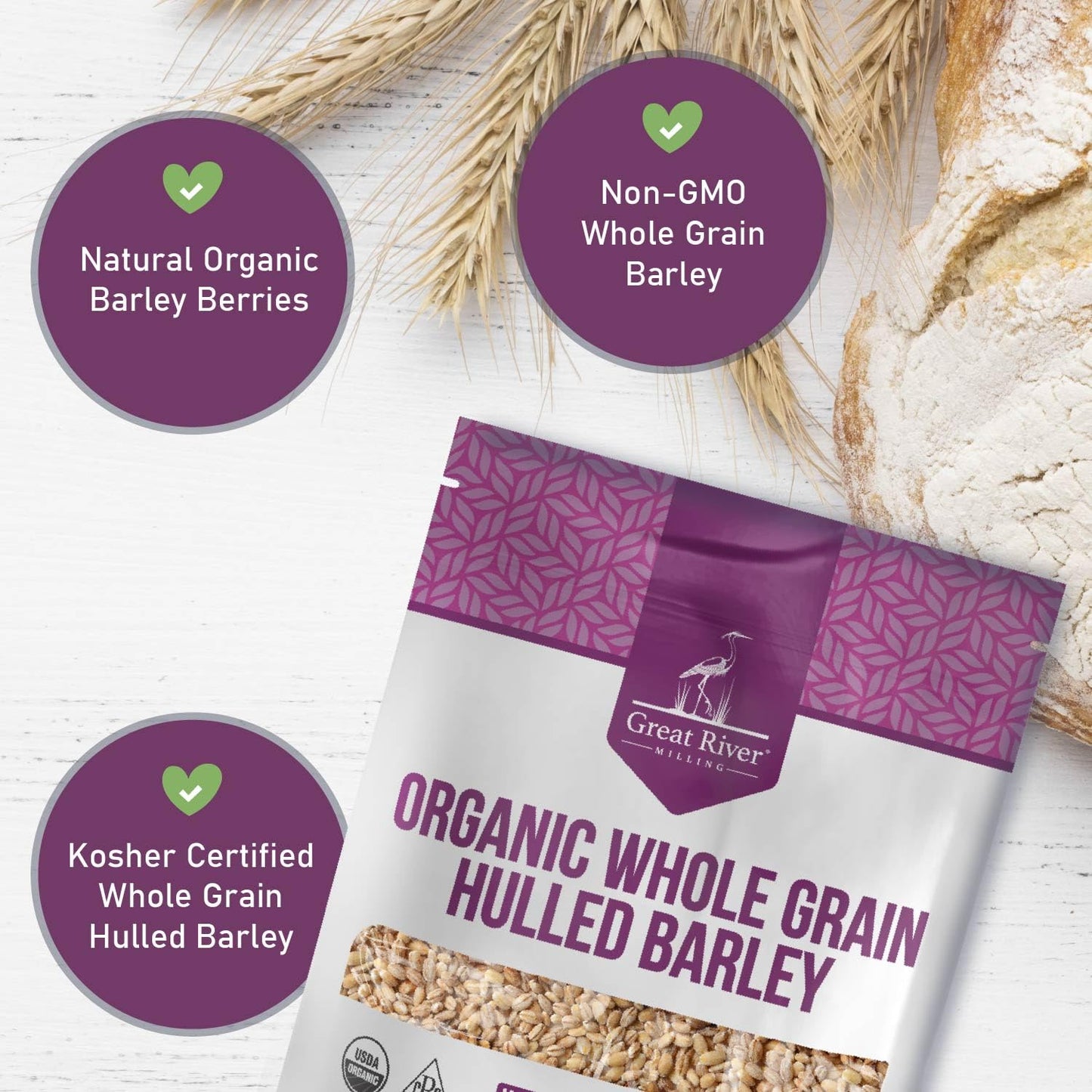 Great River Milling Organic Hulled Barley package on a light wooden surface, surrounded by wheat stalks and a rustic loaf of bread, with purple accents and product details