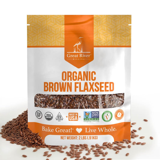 A 25 lb orange and white bag of Great River Milling Organic Brown Flaxseed, adorned with certification logos, sits upright with some seeds scattered around it