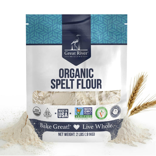 A 2 lb bag of Organic Spelt Flour featuring certification logos, illustrations of wheat stalks, and a touch of spilled flour around the package. This ancient grain flour comes in a blue and white bag displaying product details—an ideal choice for anyone looking for premium organic spelt flour.