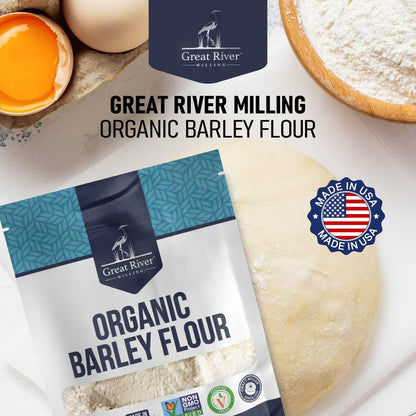 Great River Milling Organic Barley Flour package is displayed on a table with a dough ball, flour, and an egg. The package highlights its organic and non-GMO certifications
