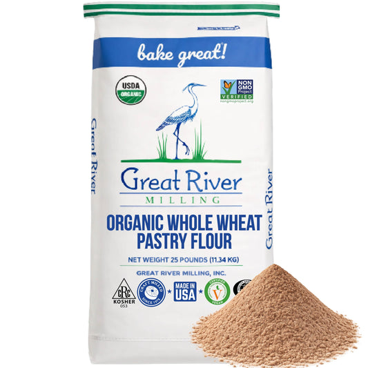 A 25 lb bag of Great River Milling Organic Whole Wheat Pastry Flour, featuring a crane illustration and certification logos, with a mound of flour in the foreground
