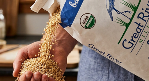 Hand holding a bag of small tan grains labeled Great River, with a circular USA stamp and blue writing